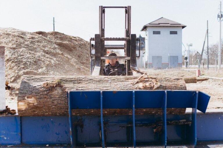 Man driving a forklift carrying logs at an outdoor industrial site.